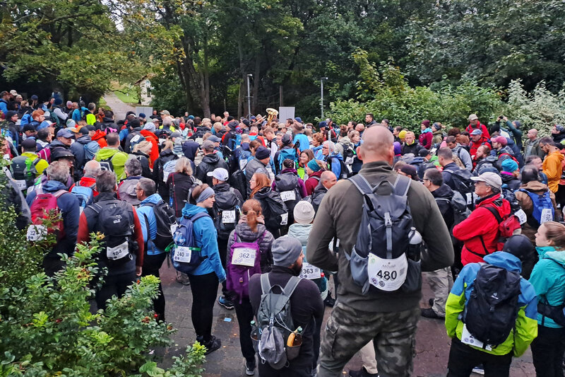Große Gruppe von Wandernden versammelt sich mit Rucksäcken und Startnummern bei einer Outdoor-Veranstaltung im Grünen.