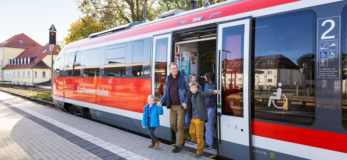Eine Familie mit mehreren Kindern steigt fröhlich aus einem Zug der Kurhessenbahn an einem sonnigen Bahnhof aus.