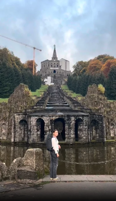 Eine Frau mit einem schwarzen Rucksack steht vor einer historischen Wasseranlage mit Bögen und Kaskaden. Im Hintergrund erhebt sich ein monumentales Bauwerk mit einer Statue an der Spitze, umgeben von grünen Bäumen mit herbstlich gefärbtem Laub. 