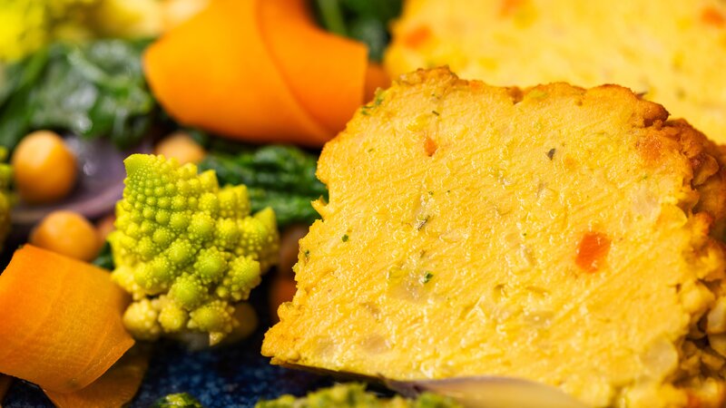 Ein saftig gebackener Kichererbsenbraten, serviert mit buntem Gemüse wie Romanesco, Karottenstreifen und Spinat.