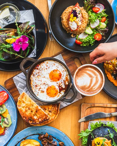Ein reich gedeckter Tisch mit verschiedenen bunten Speisen, darunter ein Topf mit Eiern in Tomatensoße, frische Salate und geröstetes Brot. Eine Hand greift nach einer Tasse Cappuccino mit Latte Art.