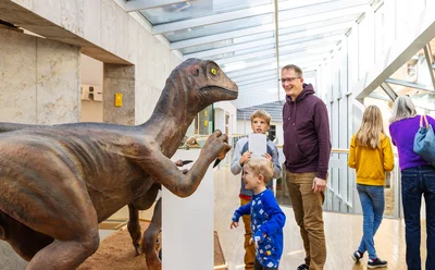 Eine Gruppe von Menschen, darunter Kinder und Erwachsene, befindet sich in einem Museum mit einem überdachten Glasgang. Ein großes Dinosauriermodell steht auf einer Sandfläche. Ein kleiner Junge in blauer Kleidung betrachtet es neugierig, während ein Mann in lila Hoodie daneben lächelt.