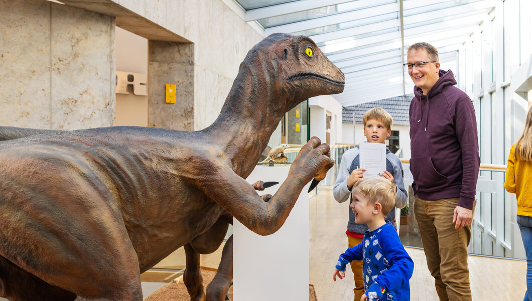 Eine Gruppe von Menschen, darunter Kinder und Erwachsene, befindet sich in einem Museum mit einem überdachten Glasgang. Ein großes Dinosauriermodell steht auf einer Sandfläche. Ein kleiner Junge in blauer Kleidung betrachtet es neugierig, während ein Mann in lila Hoodie daneben lächelt.