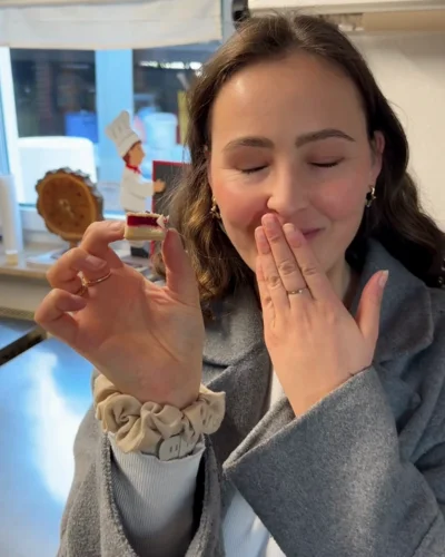 Eine junge Frau mit langen braunen Haaren und goldenen Ohrringen steht in einer Küche. Sie trägt einen grauen Mantel und hält lächelnd ein kleines Stück Kuchen zwischen Daumen und Zeigefinger ihrer linken Hand. Mit der rechten Hand bedeckt sie leicht ihren Mund – eine Geste des Genusses.