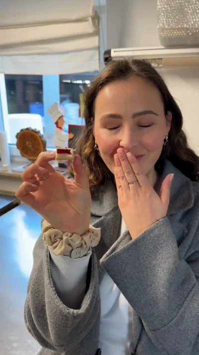 Eine junge Frau mit langen braunen Haaren und goldenen Ohrringen steht in einer Küche. Sie trägt einen grauen Mantel und hält lächelnd ein kleines Stück Kuchen zwischen Daumen und Zeigefinger ihrer linken Hand. Mit der rechten Hand bedeckt sie leicht ihren Mund – eine Geste des Genusses.