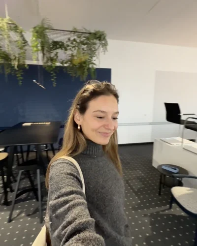 Weiblich erscheinende Person steht in einem Innenraum des Museum Thonet mit Tischen, Stühlen und Pflanzen und lächelt.