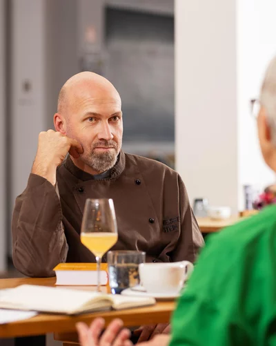 Ein Mann in einer dunklen Kochjacke sitzt an einem Tisch in einem modernen Café oder Restaurant. Er lehnt den Kopf auf eine Hand und hört einer Frau mit kurzen grauen Haaren und grünem Oberteil zu, die mit ihm spricht. Auf dem Tisch stehen Getränke, eine Tasse Kaffee und ein aufgeschlagenes Notizbuch.