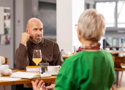 Ein Mann in einer dunklen Kochjacke sitzt an einem Tisch in einem modernen Café oder Restaurant. Er lehnt den Kopf auf eine Hand und hört einer Frau mit kurzen grauen Haaren und grünem Oberteil zu, die mit ihm spricht. Auf dem Tisch stehen Getränke, eine Tasse Kaffee und ein aufgeschlagenes Notizbuch.