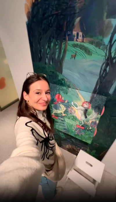 Eine junge Frau mit langen dunklen Haaren und einem hellen Pullover macht ein Selfie vor einer großen, illustrierten Wandtafel in einer Ausstellung. Das Bild zeigt eine Waldszene mit einem Haus, einem Reh und Fabelwesen mit roten Hüten. 