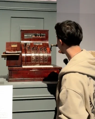 Ein junger Mann in einer beigen Kapuzenjacke steht vor einer alten hölzernen Registrierkasse in einem Museum. Er betrachtet sie aufmerksam und greift mit einer Hand an die seitliche Kurbel. Die Registrierkasse hat mehrere Hebel und eine Anzeige mit der Währung ‚Mark Pfennig‘. Neben der Ausstellung ist eine Infotafel angebracht.