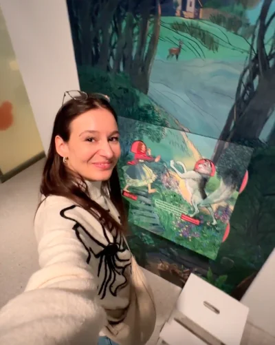 Eine junge Frau mit langen dunklen Haaren und einem hellen Pullover macht ein Selfie vor einer großen, illustrierten Wandtafel in einer Ausstellung. Das Bild zeigt eine Waldszene mit einem Haus, einem Reh und Fabelwesen mit roten Hüten. 