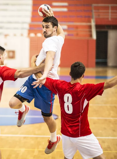 Ein Handballspieler in einem weißen Trikot und blauen Shorts springt in die Luft, um den Ball zu werfen. Zwei gegnerische Spieler in roten Trikots versuchen, ihn zu blockieren. Das Bild ist aus der Perspektive des Tores aufgenommen.