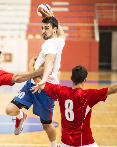 Ein Handballspieler in einem weißen Trikot und blauen Shorts springt in die Luft, um den Ball zu werfen. Zwei gegnerische Spieler in roten Trikots versuchen, ihn zu blockieren. Das Bild ist aus der Perspektive des Tores aufgenommen.