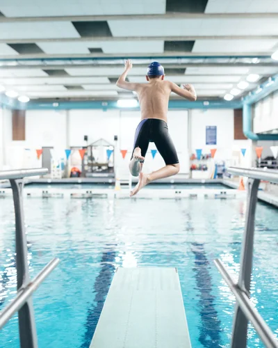 Kind springt in einem Hallenbad von einem Sprungbrett ins Wasser, das Becken und die Umgebung sind hell beleuchtet.