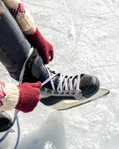 Nahaufnahme einer Person, die auf einer Eisfläche sitzt oder kniet und ihre schwarzen Schlittschuhe mit weißen Schnürsenkeln bindet. Die Person trägt rote Handschuhe und einen Strickpullover mit Muster. Der Untergrund ist eine glatte Eisfläche.