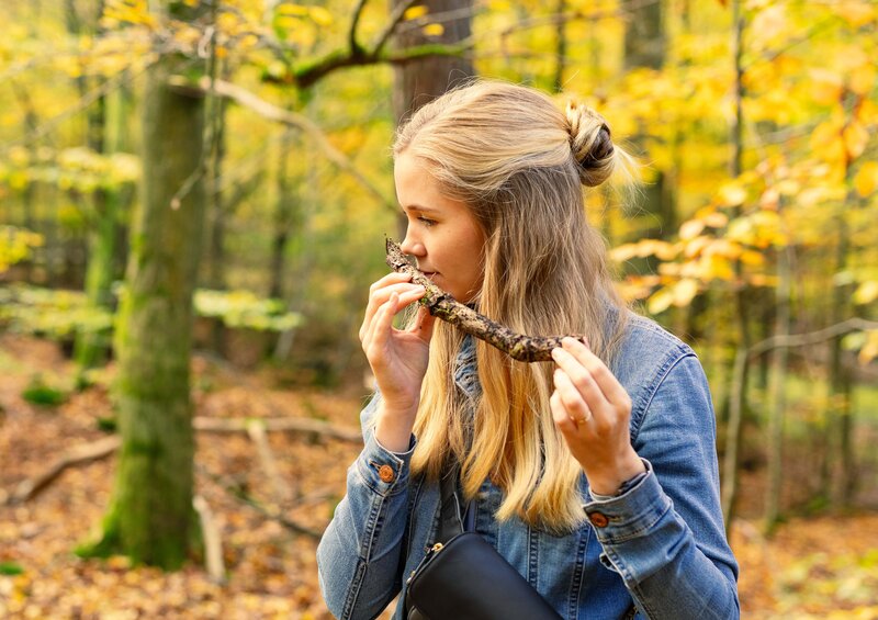 Junge Frau riecht achtsam an einem Stück Rinde im herbstlichen Wald, während im Hintergrund ein Mann ebenfalls die Natur betrachtet.