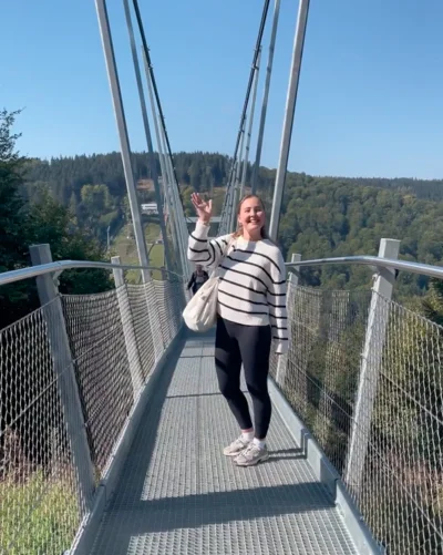 Eine Frau steht auf einer Hängebrücke aus Metall und lächelt in die Kamera. Sie trägt eine schwarz-weiß gestreifte Jacke, schwarze Leggings und helle Sneaker. Mit einer Hand winkt sie in die Kamera. Im Hintergrund sind bewaldete Hügel und ein blauer Himmel zu sehen.