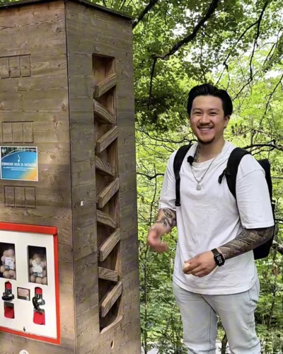 Ein junger Mann mit Rucksack lächelt in die Kamera, während er neben einer hölzernen Informationstafel im Wald steht. Im Hintergrund leuchten frische grüne Blätter.