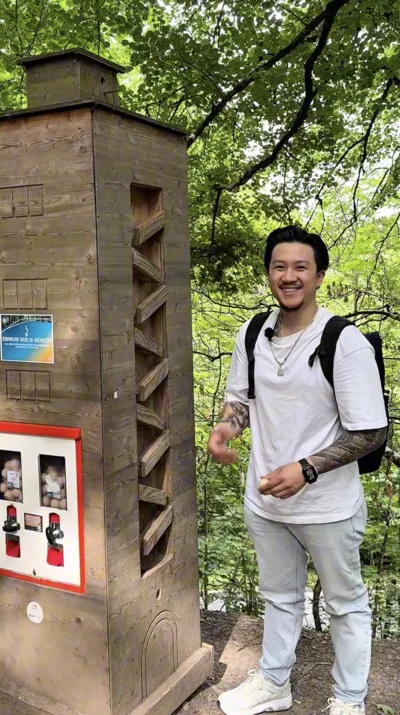 Ein junger Mann mit Rucksack lächelt in die Kamera, während er neben einer hölzernen Informationstafel im Wald steht. Im Hintergrund leuchten frische grüne Blätter.
