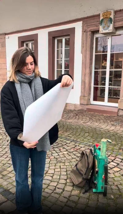 Eine junge Frau mit dunkler Jacke, grauem Schal und blauen Jeans steht auf einem gepflasterten Platz und hält ein großes, aufgerolltes Papier in den Händen. Neben ihr auf dem Boden liegen ein Rucksack und eine grüne Holzkiste mit verschiedenen Werkzeugen. Im Hintergrund ist ein historisches Gebäude mit weißen Fenstern und einem Wappen über der Tür zu sehen.