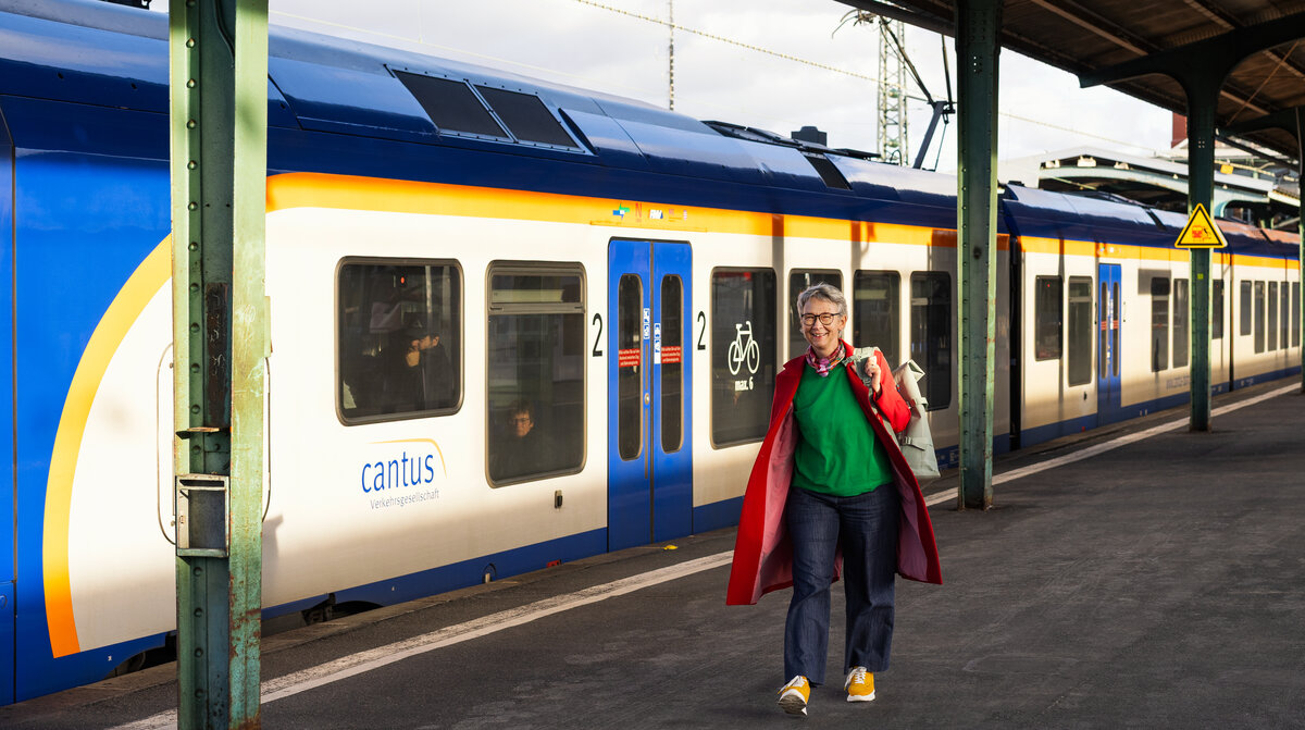 Eine Frau steigt lächelnd aus einem blauen Regionalzug der Cantus Verkehrsgesellschaft an einem Bahnsteig mit Überdachung.