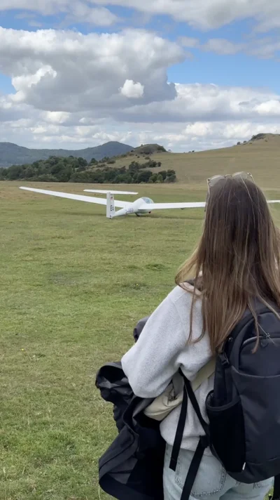 Eine junge Frau mit Rucksack blickt auf ein Segelflugzeug, das von einer offenen Wiese im Mittelgebirge abhebt. Ruhige Landschaft, weite Aussicht und ein Moment zum Staunen