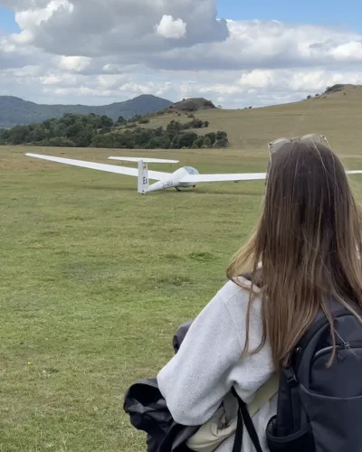 Eine junge Frau mit Rucksack blickt auf ein Segelflugzeug, das von einer offenen Wiese im Mittelgebirge abhebt. Ruhige Landschaft, weite Aussicht und ein Moment zum Staunen
