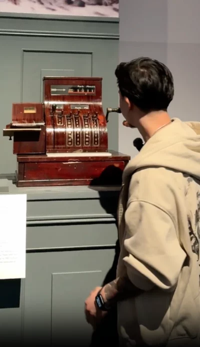 Ein junger Mann in einer beigen Kapuzenjacke steht vor einer alten hölzernen Registrierkasse in einem Museum. Er betrachtet sie aufmerksam und greift mit einer Hand an die seitliche Kurbel. Die Registrierkasse hat mehrere Hebel und eine Anzeige mit der Währung ‚Mark Pfennig‘. Neben der Ausstellung ist eine Infotafel angebracht.