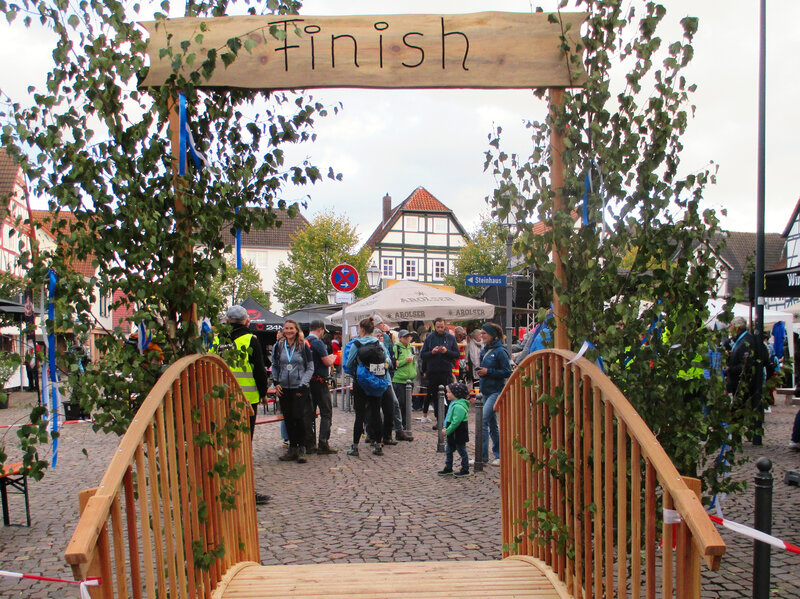 Ein dekorativer Zielbogen mit der Aufschrift „Finish“ steht auf einem Stadtplatz, wo Wandernde gerade das Ziel erreichen. Umgeben sind sie von Fachwerkhäusern, Besuchern und Marktständen.