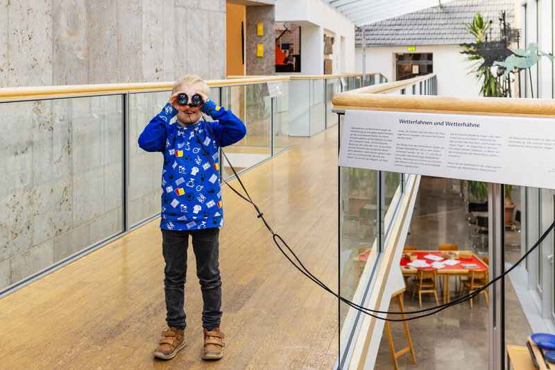 Ein blondes Kind steht in einem hellen Museumsflur mit Glasdach und schaut durch ein Fernglas.