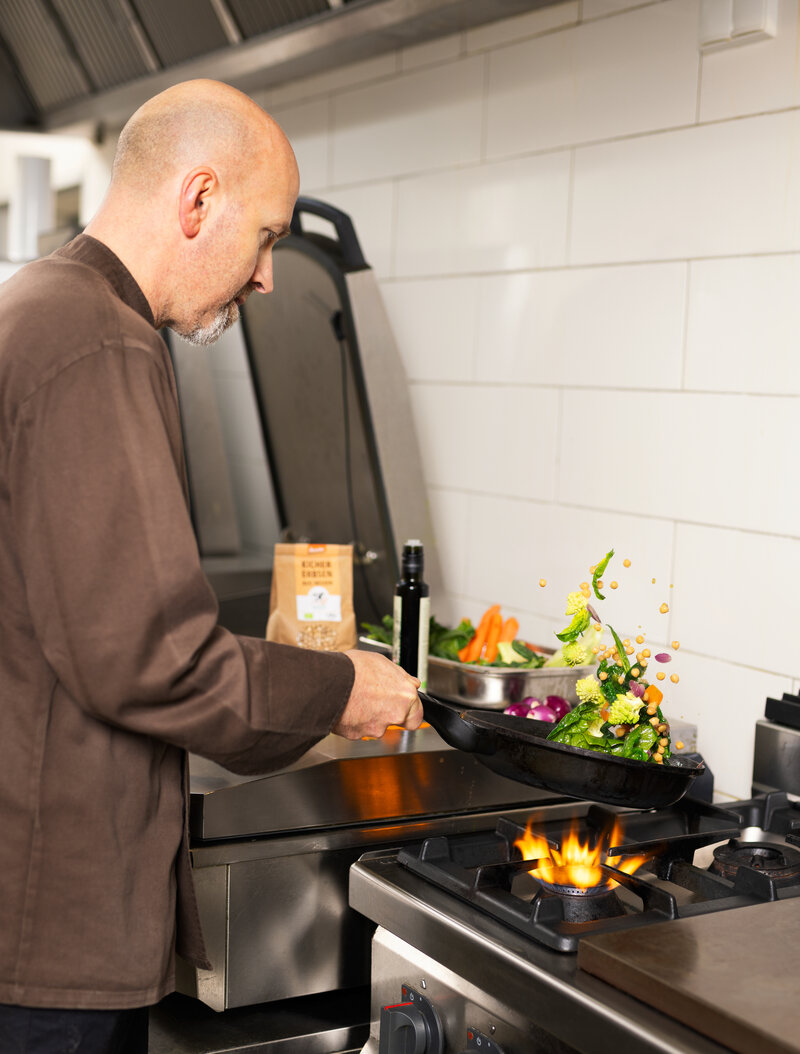 Ein Koch wirbelt frisches Gemüse in einer Pfanne über einer offenen Gasflamme in einer professionellen Küche.