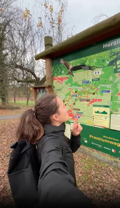 Eine Frau mit dunkler Jacke und Rucksack steht vor einer großen grünen Informationstafel in einem Park oder Naturgebiet. Sie zeigt mit dem Finger auf die Karte und blickt konzentriert darauf. Im Hintergrund sind kahle Bäume und ein Laubboden zu sehen.