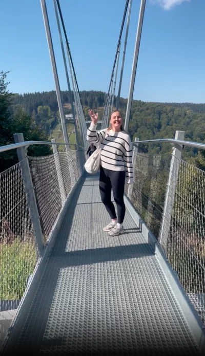 Eine Frau steht auf einer Hängebrücke aus Metall und lächelt in die Kamera. Sie trägt eine schwarz-weiß gestreifte Jacke, schwarze Leggings und helle Sneaker. Mit einer Hand winkt sie in die Kamera. Im Hintergrund sind bewaldete Hügel und ein blauer Himmel zu sehen.