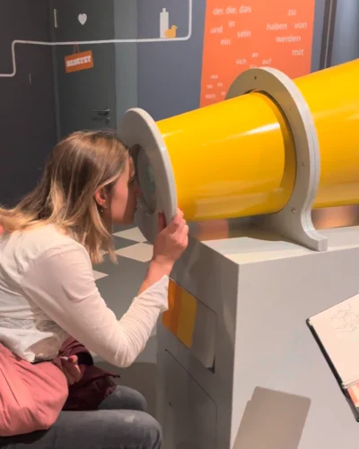 Eine Frau mit heller Kleidung sitzt auf einem Hocker und schaut durch ein großes gelbes Rohr in einer interaktiven Ausstellung. Im Hintergrund sind eine Tür mit einem Herzsymbol und eine Wand mit deutschen Wörtern in orangefarbenem Design zu sehen.
