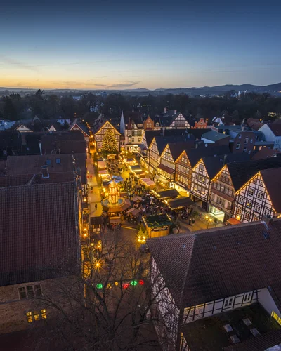 Luftaufnahme eines festlich beleuchteten Weihnachtsmarktes in einer historischen Altstadt mit Fachwerkhäusern bei Dämmerung. Zahlreiche Marktstände und Lichter sorgen für eine stimmungsvolle Atmosphäre.