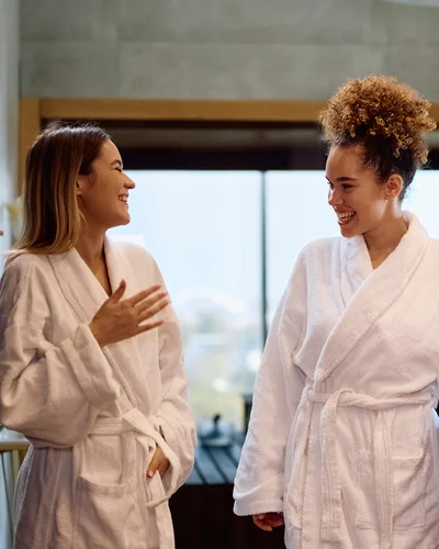 Zwei weiblich erscheinende Personen im Bademantel stehen in einem Wellnessbereich und unterhalten sich lächelnd