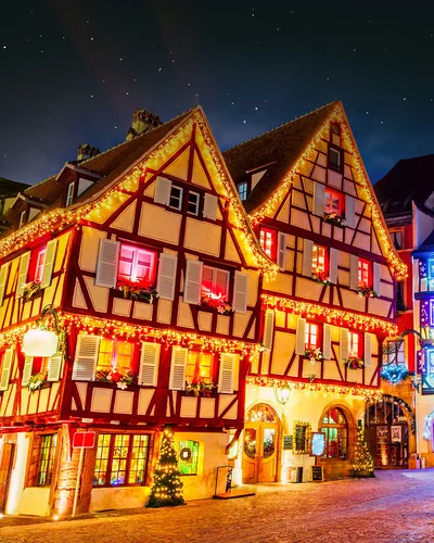 Zwei traditionelle Fachwerkhäuser sind festlich mit Lichterketten und Weihnachtsdekoration geschmückt. Die Fenster leuchten warm, und es sind rote Schleifen sowie festliche Ornamente zu sehen. Der Nachthimmel ist dunkel, während die Beleuchtung die Fassaden in ein stimmungsvolles Licht taucht. Weitere geschmückte Gebäude und Lichter sind im Hintergrund erkennbar.