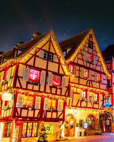 Zwei traditionelle Fachwerkhäuser sind festlich mit Lichterketten und Weihnachtsdekoration geschmückt. Die Fenster leuchten warm, und es sind rote Schleifen sowie festliche Ornamente zu sehen. Der Nachthimmel ist dunkel, während die Beleuchtung die Fassaden in ein stimmungsvolles Licht taucht. Weitere geschmückte Gebäude und Lichter sind im Hintergrund erkennbar.
