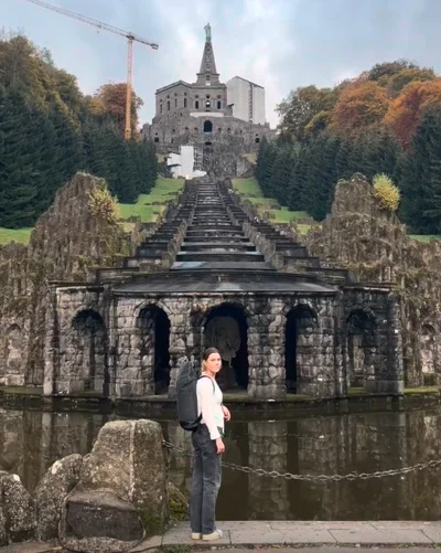 Eine Frau mit einem schwarzen Rucksack steht vor einer historischen Wasseranlage mit Bögen und Kaskaden. Im Hintergrund erhebt sich ein monumentales Bauwerk mit einer Statue an der Spitze, umgeben von grünen Bäumen mit herbstlich gefärbtem Laub. 