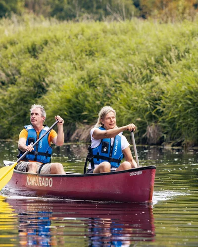Zwei Personen paddeln in einem roten Kanu auf einem ruhigen Fluss, umgeben von grüner Ufervegetation