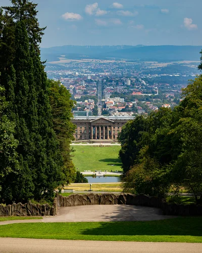 Panoramablick auf das Schloss Wilhelmshöhe in Kassel, eingerahmt von Bäumen und mit Blick auf die Stadt im Hintergrund.