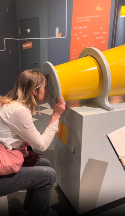 Eine Frau mit heller Kleidung sitzt auf einem Hocker und schaut durch ein großes gelbes Rohr in einer interaktiven Ausstellung. Im Hintergrund sind eine Tür mit einem Herzsymbol und eine Wand mit deutschen Wörtern in orangefarbenem Design zu sehen.
