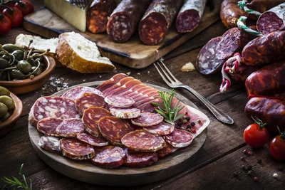 Auf einem Holzteller angerichtete Scheiben von Ahle Wurscht und luftgetrocknetem Schinken, umgeben von Brot, Oliven, Kapern und Tomaten