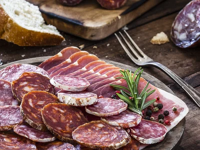 Auf einem Holzteller angerichtete Scheiben von Ahle Wurscht und luftgetrocknetem Schinken, umgeben von Brot, Oliven, Kapern und Tomaten