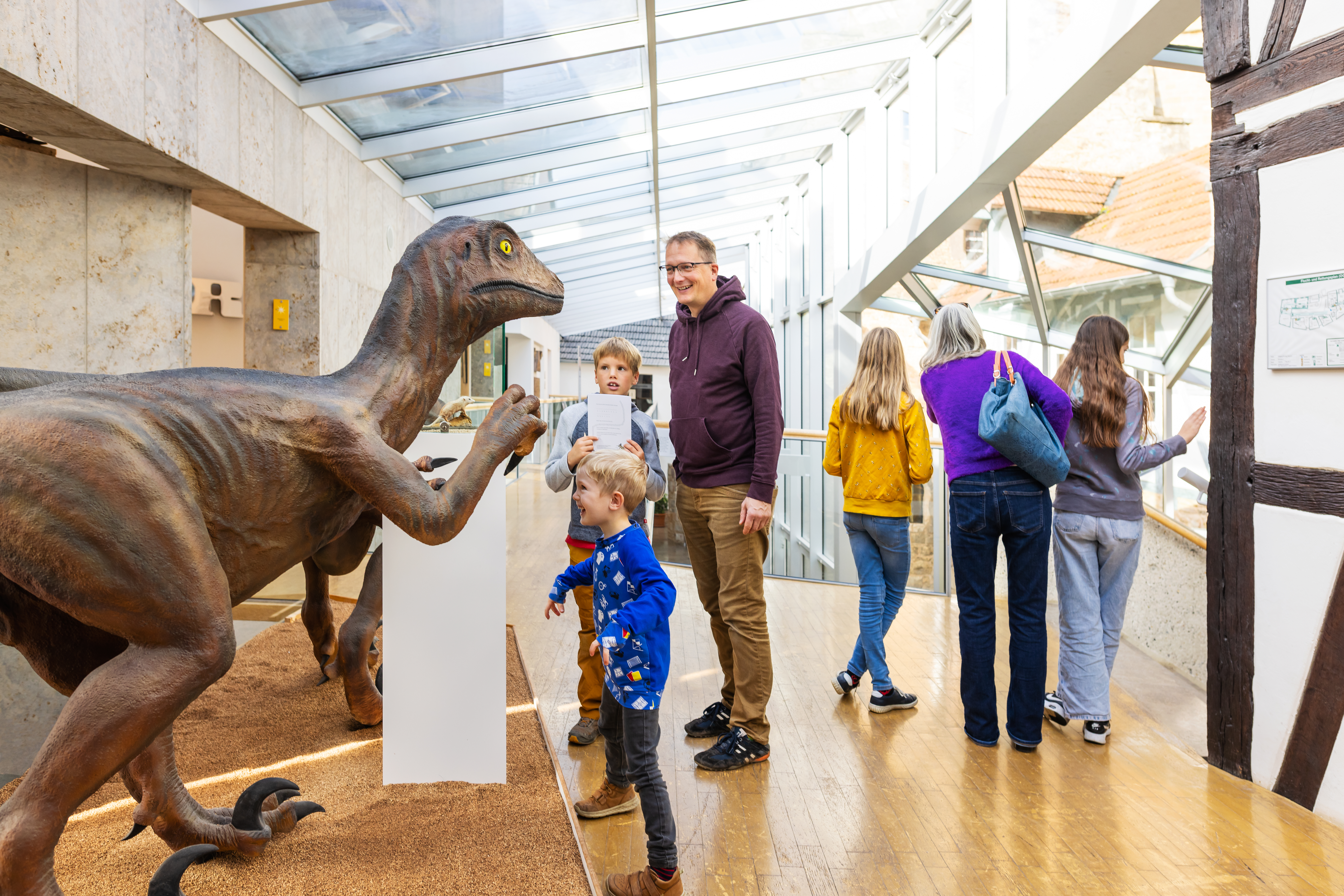 Eine Gruppe von Menschen, darunter Kinder und Erwachsene, befindet sich in einem Museum mit einem überdachten Glasgang. Ein großes Dinosauriermodell steht auf einer Sandfläche. Ein kleiner Junge in blauer Kleidung betrachtet es neugierig, während ein Mann in lila Hoodie daneben lächelt.