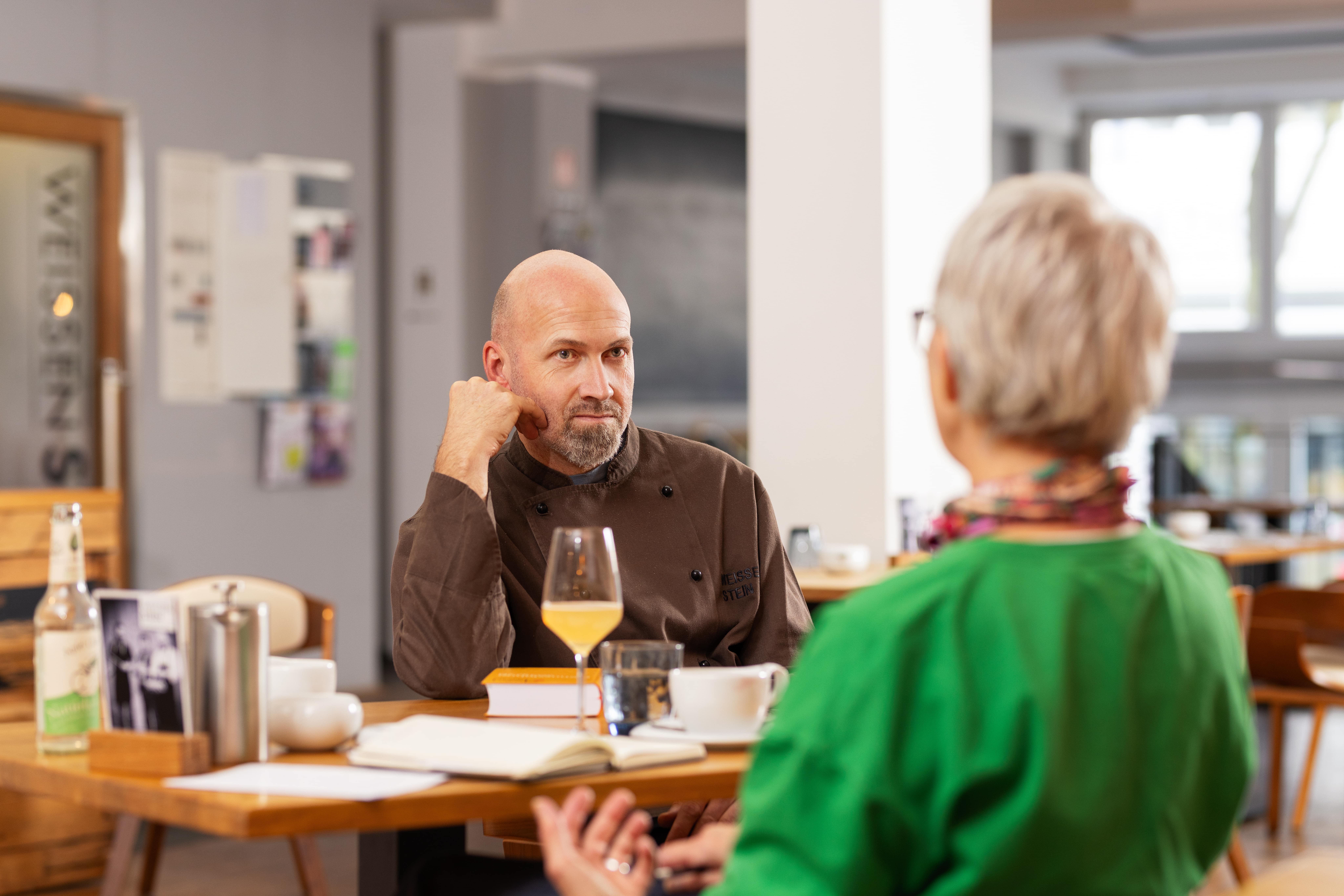 Ein Mann in einer dunklen Kochjacke sitzt an einem Tisch in einem modernen Café oder Restaurant. Er lehnt den Kopf auf eine Hand und hört einer Frau mit kurzen grauen Haaren und grünem Oberteil zu, die mit ihm spricht. Auf dem Tisch stehen Getränke, eine Tasse Kaffee und ein aufgeschlagenes Notizbuch.